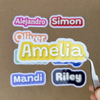 Tweezers hold a bright yellow custom name sticker for "Amelia" in a bold font, showcasing the large 4-inch width against other colorful name decals in the background.