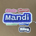 A close-up scale shot of a large 4-inch purple name sticker for "Mandi" being held by tweezers, demonstrating its size relative to other personalized vinyl decals.