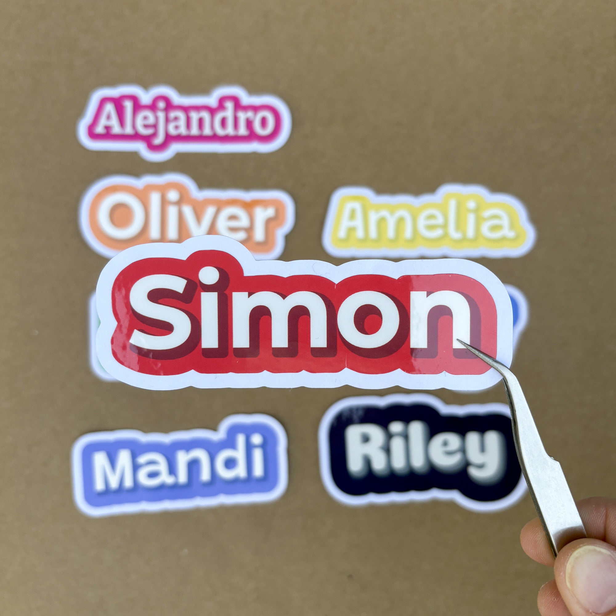 A hand holding tweezers points to a bold red 4-inch wide custom name sticker reading "Simon," illustrating the high-quality precision-cut vinyl.