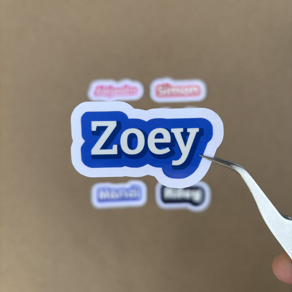Tweezers holding a 2-inch wide blue name sticker for "Zoey," demonstrating its smaller, accessory-friendly size against a neutral background.