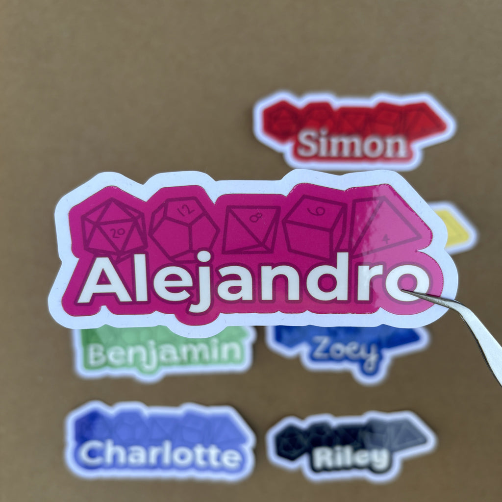 Tweezers hold a vibrant pink 1.5-inch tall custom name sticker for "Alejandro" with a d20 and d12 dice motif in the background.