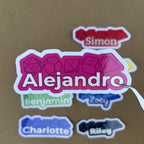 Tweezers hold a vibrant pink 1.5-inch tall custom name sticker for "Alejandro" with a d20 and d12 dice motif in the background.