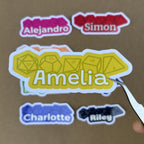 A close-up scale shot of a yellow custom name sticker for "Amelia" featuring a tabletop RPG dice design, held by silver tweezers.