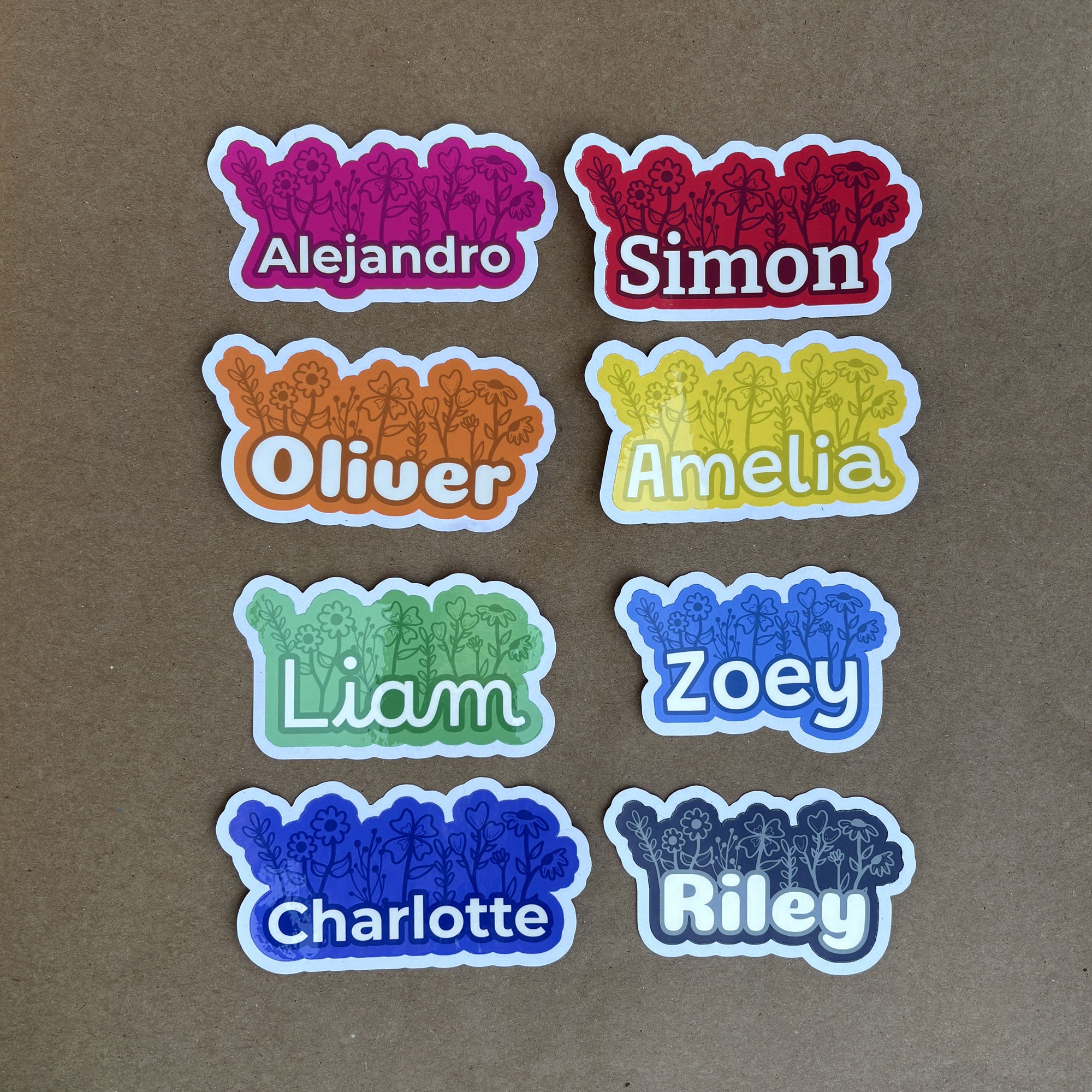 A grid display of eight custom floral name stickers showcasing the full color palette: Pink (Alejandro), Red (Simon), Orange (Oliver), Yellow (Amelia), Green (Liam), Blue (Zoey), Dark Blue (Charlotte), and Grey (Riley).
