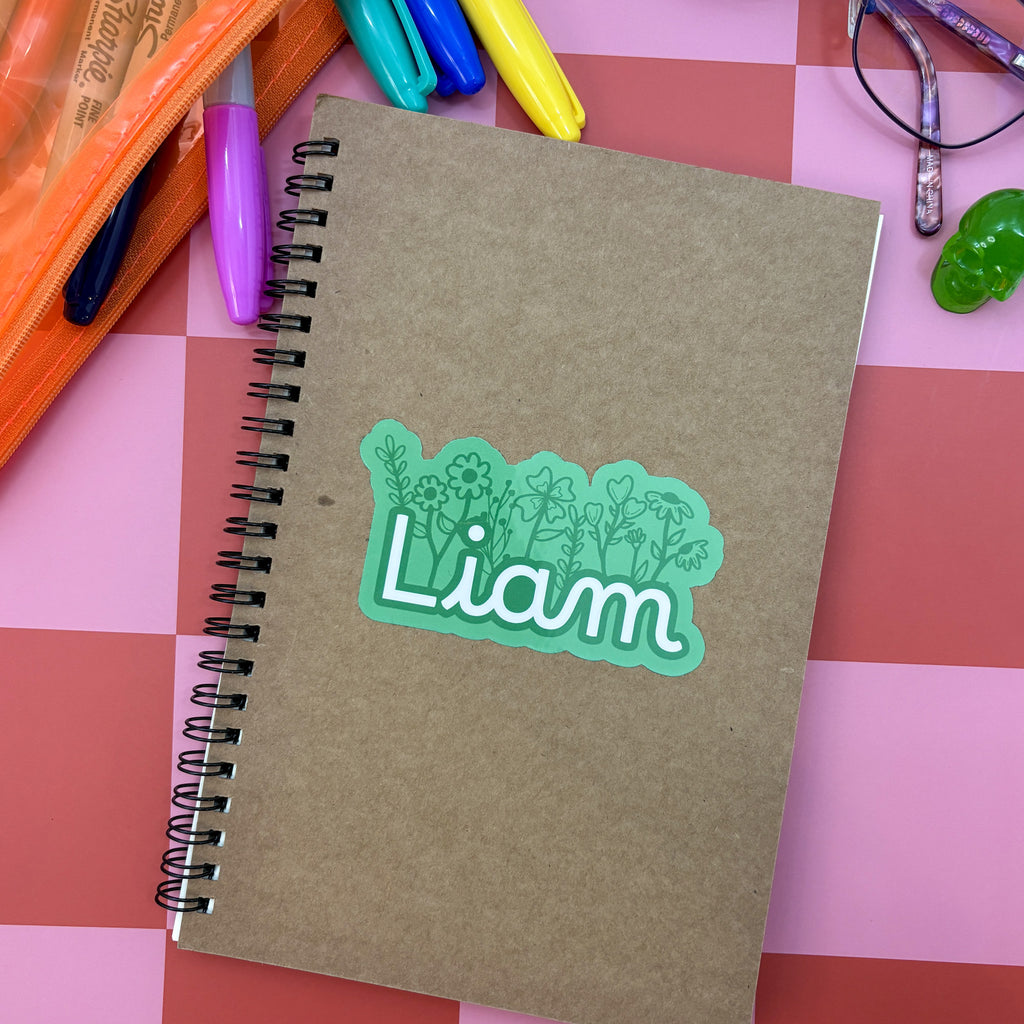 A custom 2-inch green vinyl name sticker reading "Liam" in a white script font, featuring a hand-drawn floral background. The sticker is placed on a brown kraft paper spiral notebook surrounded by colorful markers and a green skull.