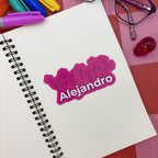 A personalized pink vinyl sticker with the name "Alejandro" in bold white text against a floral motif. The sticker is centered on a clean white page of a spiral sketchbook next to pink markers and glasses.