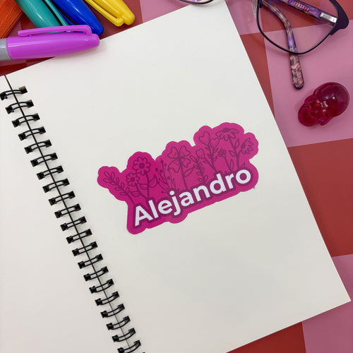 A personalized pink vinyl sticker with the name "Alejandro" in bold white text against a floral motif. The sticker is centered on a clean white page of a spiral sketchbook next to pink markers and glasses.