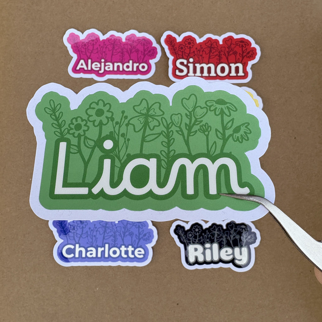 A close-up scale shot showing a large green "Liam" floral sticker being held by tweezers. Various smaller name stickers in red, pink, blue, and black are displayed in the background to show size comparison.