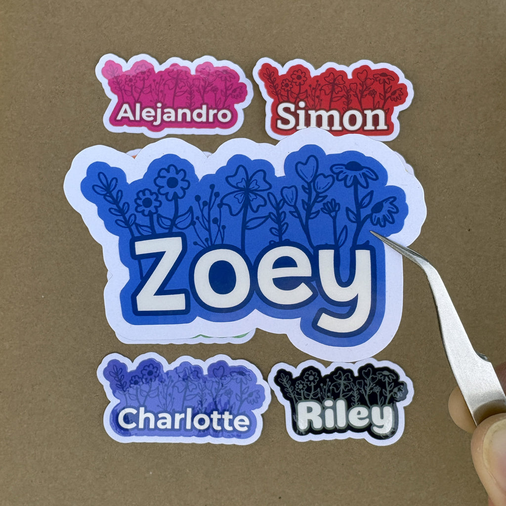 A hand holding silver tweezers points to a large blue custom name sticker for "Zoey." Smaller stickers for "Alejandro," "Simon," "Charlotte," and "Riley" are shown above and below to demonstrate the 2-inch height scale.